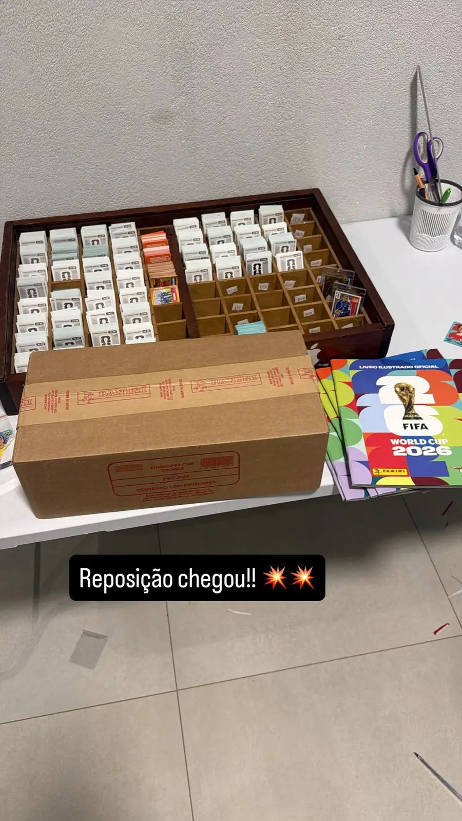 Caixa lacrada da Panini com 1.000 envelopes ao lado de álbuns oficiais